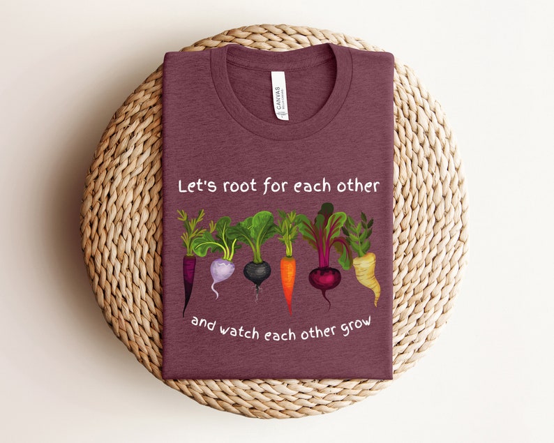 May include: A maroon t-shirt with the text "Let's root for each other and watch each other grow" printed in white. The text is surrounded by a graphic of five different root vegetables.