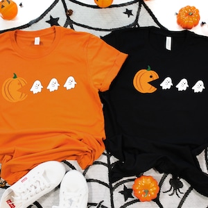Może przedstawiać: Dwie koszulki z motywem Halloween. Jedna koszulka jest pomarańczowa z dynią, kt&oacute;ra zjada duchy. Druga koszulka jest czarna z duchem, kt&oacute;ry zjada dynię.