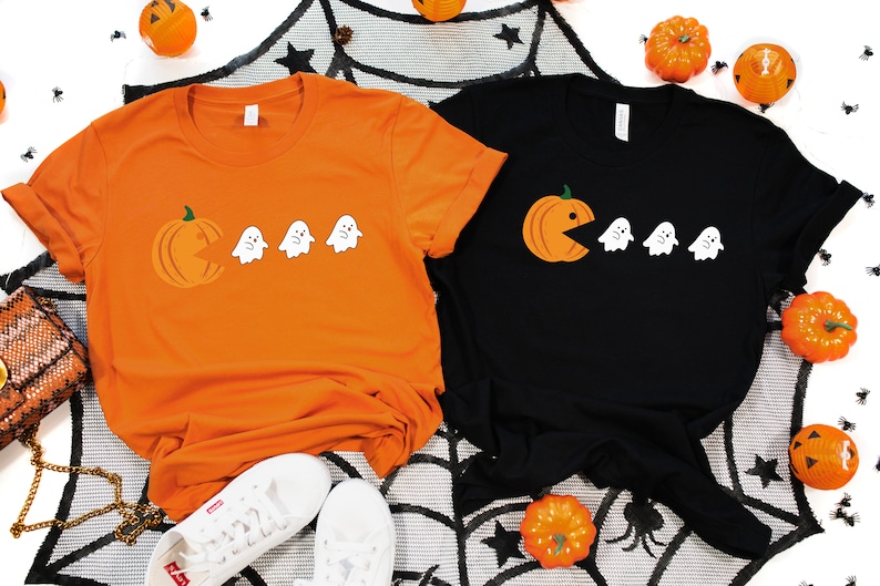 Może przedstawiać: Dwie koszulki z motywem Halloween. Jedna koszulka jest pomarańczowa z dynią, kt&oacute;ra zjada duchy. Druga koszulka jest czarna z duchem, kt&oacute;ry zjada dynię.