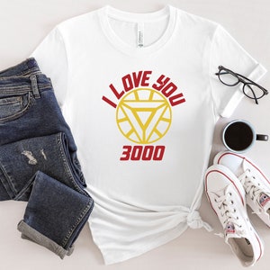 Puede incluir: Camiseta blanca con un gr&aacute;fico rojo y amarillo de la m&aacute;scara de Iron Man y el texto "I Love You 3000".