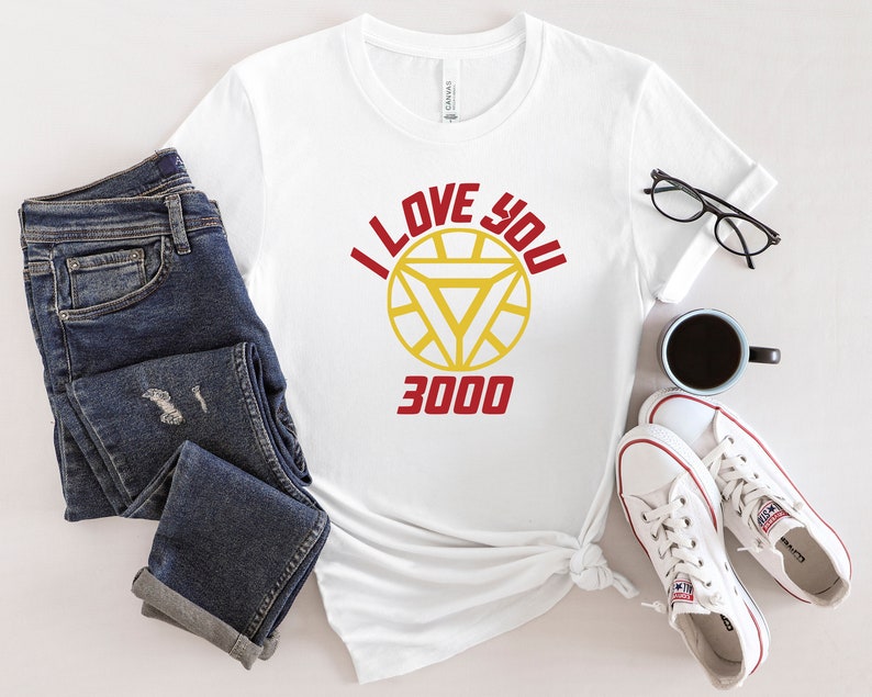 Puede incluir: Camiseta blanca con un gr&aacute;fico rojo y amarillo de la m&aacute;scara de Iron Man y el texto "I Love You 3000".