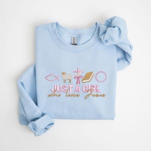 May include: Light blue sweatshirt with a pink and gold graphic that says "Just a girl who loves Jesus" with a fish, lamb, cross, ribbon, book, and wreath.