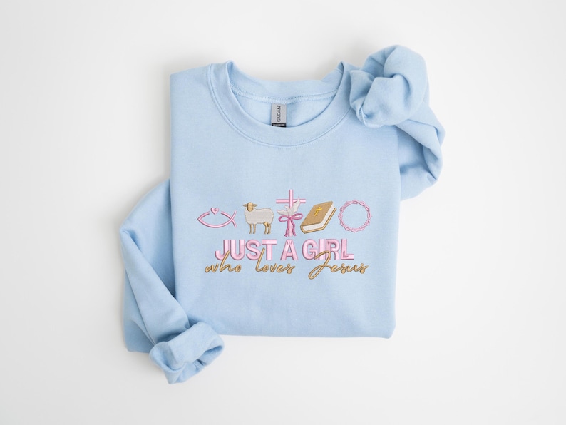 May include: Light blue sweatshirt with a pink and gold graphic that says "Just a girl who loves Jesus" with a fish, lamb, cross, ribbon, book, and wreath.