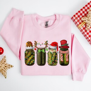 Pu&ograve; includere: Una felpa rosa con un design natalizio che presenta quattro barattoli di sottaceti con cappelli di Babbo Natale, cappelli da elfo e corna di renna. I barattoli sono decorati con luci natalizie e fiocchi.