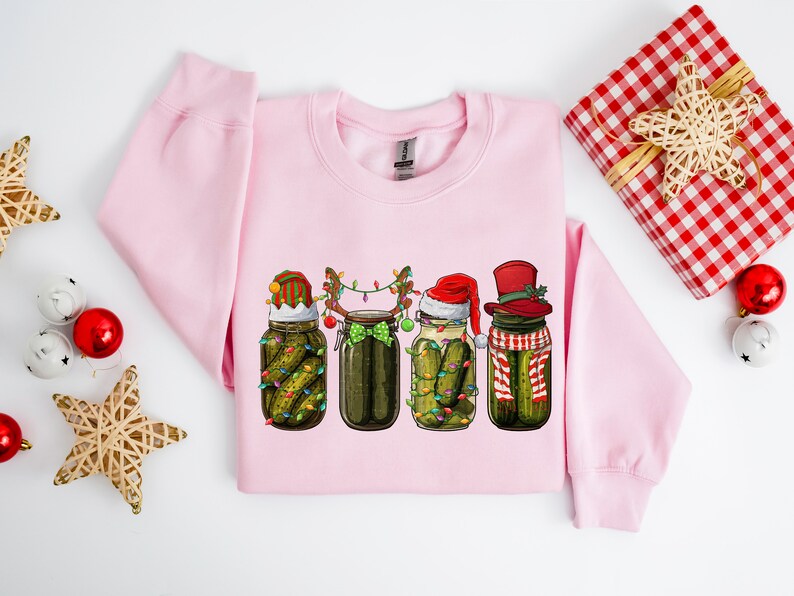 Pu&ograve; includere: Una felpa rosa con un design natalizio che presenta quattro barattoli di sottaceti con cappelli di Babbo Natale, cappelli da elfo e corna di renna. I barattoli sono decorati con luci natalizie e fiocchi.