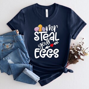 May include: A navy blue t-shirt with a white graphic that says "I'm steal your eggs" with a heart and arrow. The graphic also includes a cluster of colourful Easter eggs.