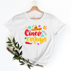 May include: White t-shirt with a colorful graphic design that says "Cinco de Mayo" with a sombrero, flowers, and a chili pepper.