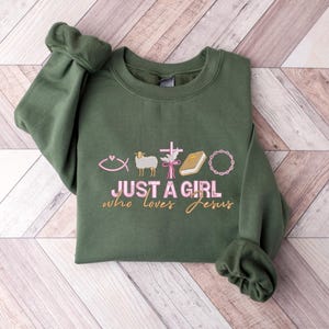 May include: A green sweatshirt with the text "Just a girl who loves Jesus" in pink lettering. The sweatshirt has embroidered Christian symbols including a fish, a lamb, a cross, a dove, a book, and a crown of thorns.