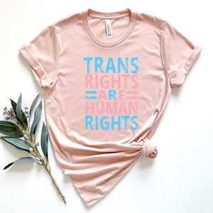 May include: A light pink t-shirt with the text "TRANS RIGHTS ARE HUMAN RIGHTS" in blue and pink lettering.