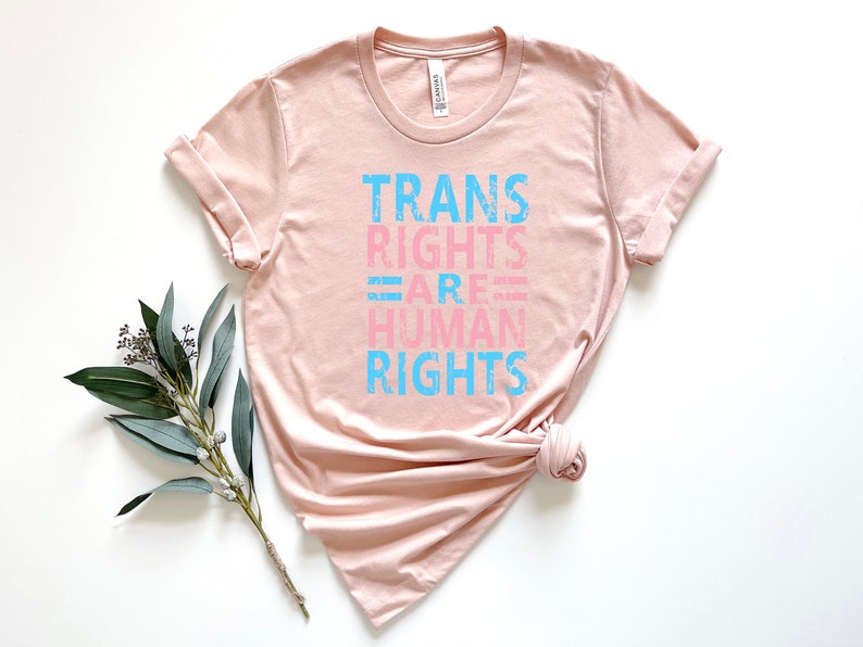 May include: A light pink t-shirt with the text "TRANS RIGHTS ARE HUMAN RIGHTS" in blue and pink lettering.