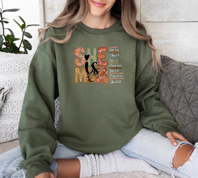 May include: Green sweatshirt with a floral design that spells out "She is Mom" with bible verses listed below. The verses are: Strong (Psalm 18:2), Able (1 Tim 1:14),  Victorious (Rom 8:37), Never Alone (Matt 28:20), Greatly Loved (Jn 3:16).