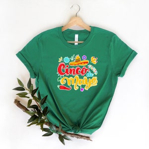 May include: Green t-shirt with a colorful graphic design that says "Cinco de Mayo" with a sombrero and flowers.