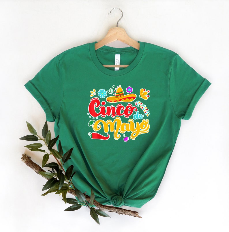 May include: Green t-shirt with a colorful graphic design that says "Cinco de Mayo" with a sombrero and flowers.
