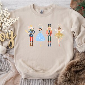 May include: A beige sweatshirt with a graphic print of a mouse, two ballerinas, and a nutcracker. The ballerinas are wearing blue and gold tutus. The mouse is wearing a crown and holding a sword. The nutcracker is wearing a red and gold uniform.