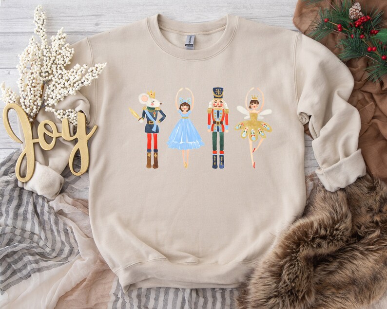 May include: A beige sweatshirt with a graphic print of a mouse, two ballerinas, and a nutcracker. The ballerinas are wearing blue and gold tutus. The mouse is wearing a crown and holding a sword. The nutcracker is wearing a red and gold uniform.