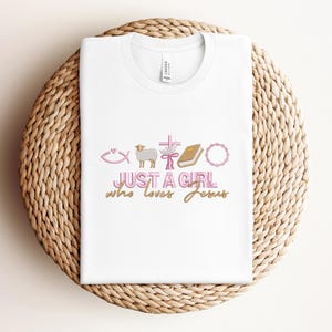 May include: White t-shirt with a pink and gold design that says "Just a girl who loves Jesus". The design includes a fish, a lamb, a cross, a bible, and a wreath.