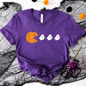 Może przedstawiać: Fioletowy T-shirt z grafiką przedstawiającą dynię jedzącą duchy. Dynia jest pomarańczowa, a duchy są białe.
