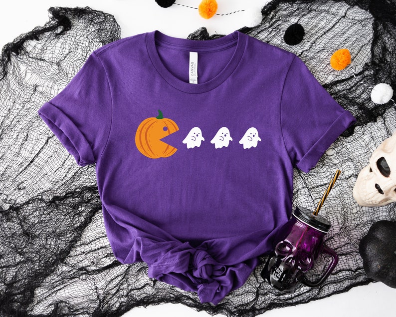 Może przedstawiać: Fioletowy T-shirt z grafiką przedstawiającą dynię jedzącą duchy. Dynia jest pomarańczowa, a duchy są białe.