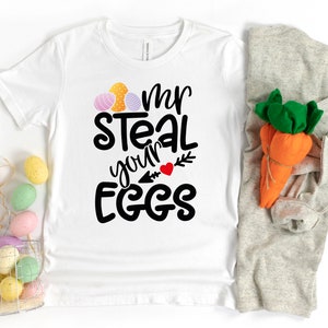 May include: White t-shirt with black text that reads "Mr Steal your Eggs" with a red heart and arrow. The shirt is decorated with three colourful Easter eggs.