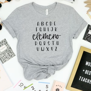 以下が含まれることがあります： アルファベットがプリントされたグレーのTシャツ。アルファベットの中で「elemeno」という単語は他の文字よりも大きなフォントサイズで書かれています。シャツは裾で結ばれています。