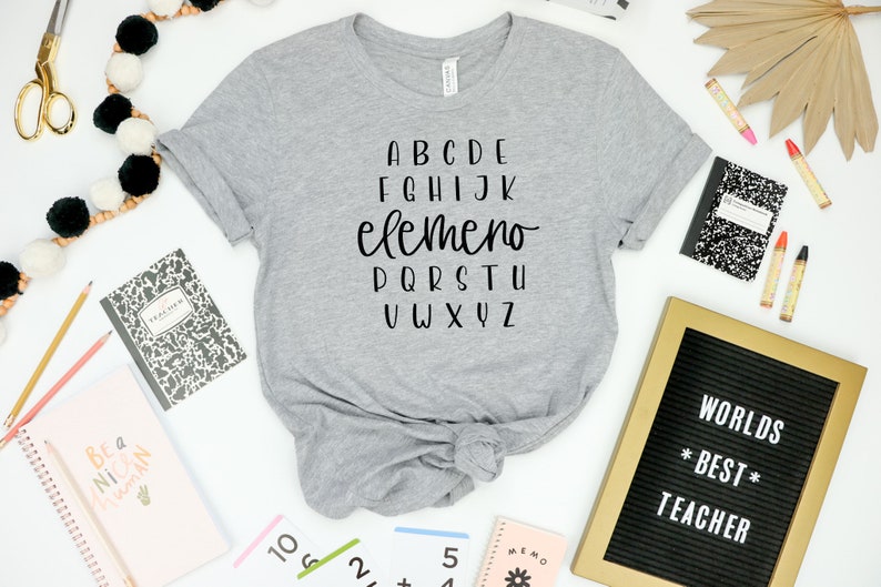以下が含まれることがあります： アルファベットがプリントされたグレーのTシャツ。アルファベットの中で「elemeno」という単語は他の文字よりも大きなフォントサイズで書かれています。シャツは裾で結ばれています。