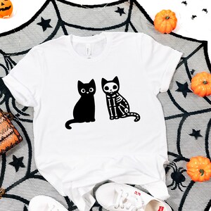 May include: White t-shirt with a black cat and a skeleton cat graphic. The shirt is on a black and white spiderweb background with orange pumpkins.