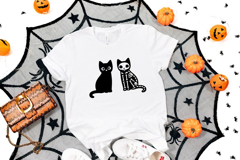 May include: White t-shirt with a black cat and a skeleton cat graphic. The shirt is on a black and white spiderweb background with orange pumpkins.
