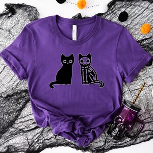 May include: Purple t-shirt with a black cat and a skeleton cat graphic. The cats are sitting side-by-side.