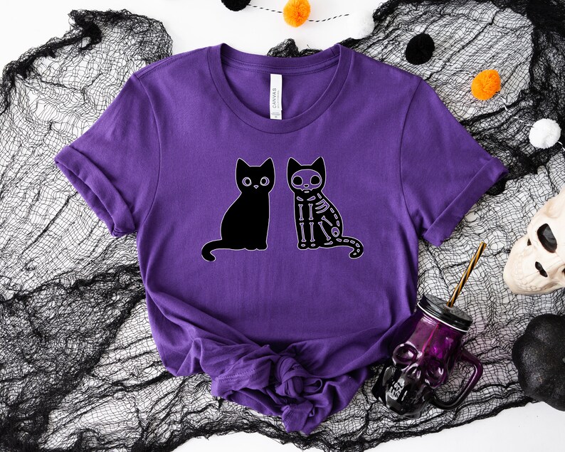 May include: Purple t-shirt with a black cat and a skeleton cat graphic. The cats are sitting side-by-side.