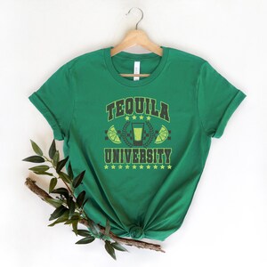 May include: Green t-shirt with the text "Tequila University" in black and yellow lettering. The design includes a shot glass with a lime wedge and stars.