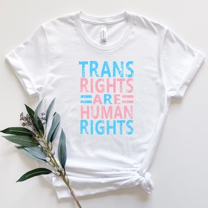 May include: White t-shirt with the text "TRANS RIGHTS ARE HUMAN RIGHTS" in blue and pink lettering.