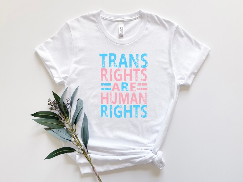 May include: White t-shirt with the text "TRANS RIGHTS ARE HUMAN RIGHTS" in blue and pink lettering.