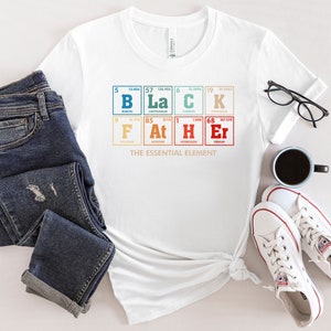 Puede incluir: Camiseta blanca con un gr&aacute;fico de la tabla peri&oacute;dica de los elementos que deletrea "BLACK" con el texto "THE ESSENTIAL ELEMENT" debajo. La camiseta se muestra sobre una superficie blanca con un par de jeans, una taza de caf&eacute; y zapatillas blancas.