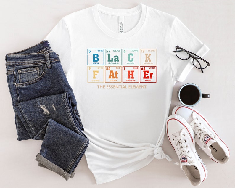 Puede incluir: Camiseta blanca con un gr&aacute;fico de la tabla peri&oacute;dica de los elementos que deletrea "BLACK" con el texto "THE ESSENTIAL ELEMENT" debajo. La camiseta se muestra sobre una superficie blanca con un par de jeans, una taza de caf&eacute; y zapatillas blancas.