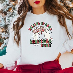 Puede incluir: Sudadera blanca con un dise&ntilde;o gr&aacute;fico verde, rojo y blanco que dice "Rolling Up Some Christmas Spirit" y representa dos manos enrollando un porro con un bast&oacute;n de caramelo dentro.