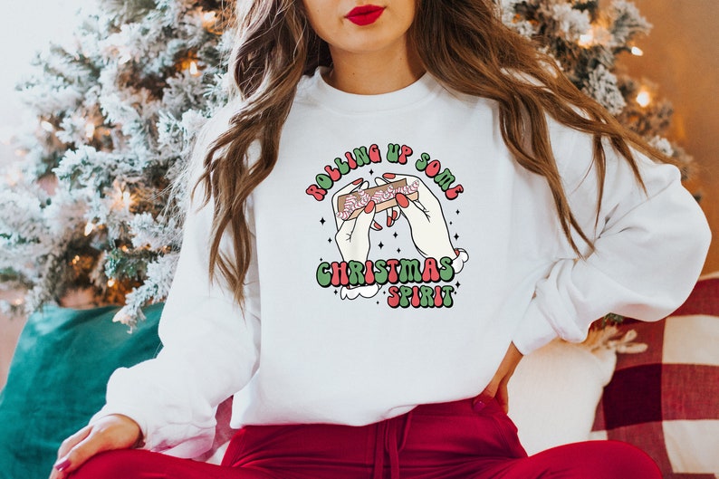 Puede incluir: Sudadera blanca con un dise&ntilde;o gr&aacute;fico verde, rojo y blanco que dice "Rolling Up Some Christmas Spirit" y representa dos manos enrollando un porro con un bast&oacute;n de caramelo dentro.