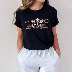 May include: Black t-shirt with a pink and gold graphic that says "Just a girl who loves Jesus" and includes Christian symbols like a fish, a cross, a lamb, a book, and a crown.