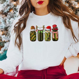 Pu&ograve; includere: Felpa bianca con un design a tema natalizio con quattro barattoli di sottaceti che indossano cappelli e accessori festivi. I barattoli sono decorati con luci e verde.