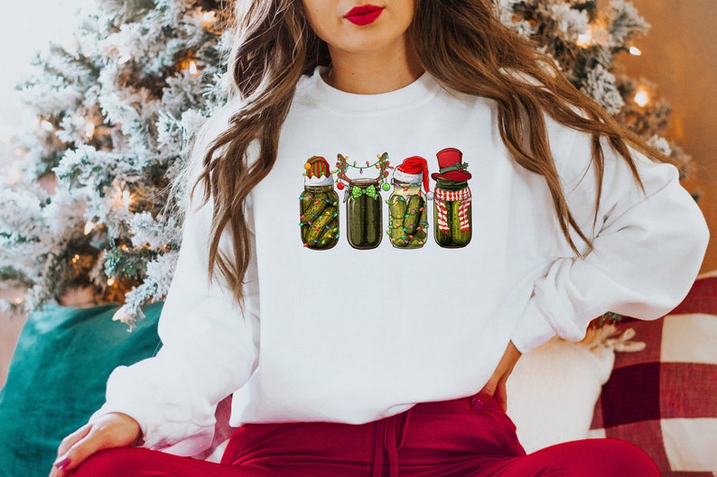 Pu&ograve; includere: Felpa bianca con un design a tema natalizio con quattro barattoli di sottaceti che indossano cappelli e accessori festivi. I barattoli sono decorati con luci e verde.
