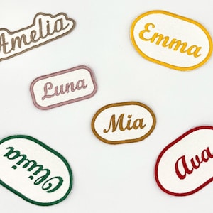 May include: Embroidered name patches in various colors. The names Amelia, Emma, Luna, Mia, Olivia, and Ava are displayed in cursive font on white patches with colored borders. These patches are ideal for personalizing clothing or accessories.