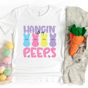 May include: White t-shirt with a pink, yellow, blue, and purple peep design and the text "Hangin' with my peeps".