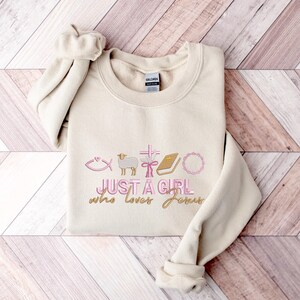 May include: A beige sweatshirt with the text "Just a girl who loves Jesus" in pink and gold lettering. The design includes a fish, a lamb, a cross, a book, and a wreath.