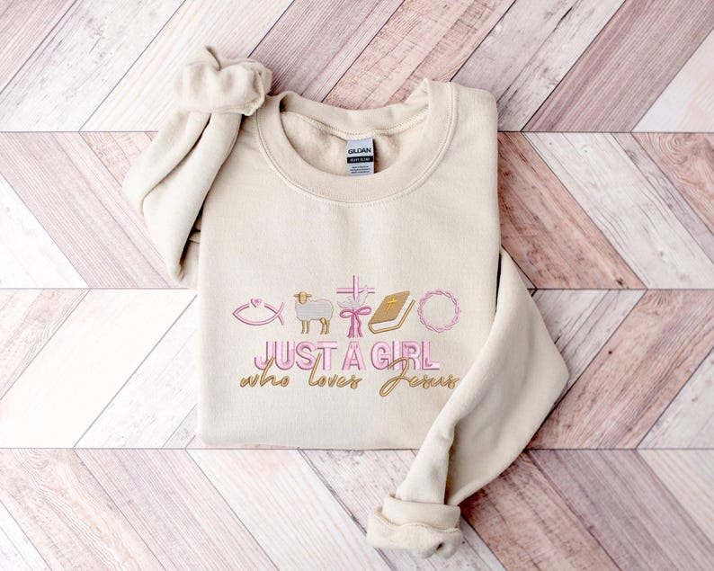 May include: A beige sweatshirt with the text "Just a girl who loves Jesus" in pink and gold lettering. The design includes a fish, a lamb, a cross, a book, and a wreath.