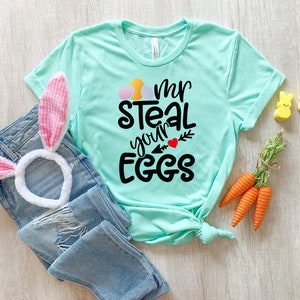 May include: A turquoise blue t-shirt with the text "I'm steal your eggs" in black lettering with a red heart and a green arrow. The shirt is folded and laid on a wooden surface with a pink bunny ear headband, carrots, and yellow chick figurines.
