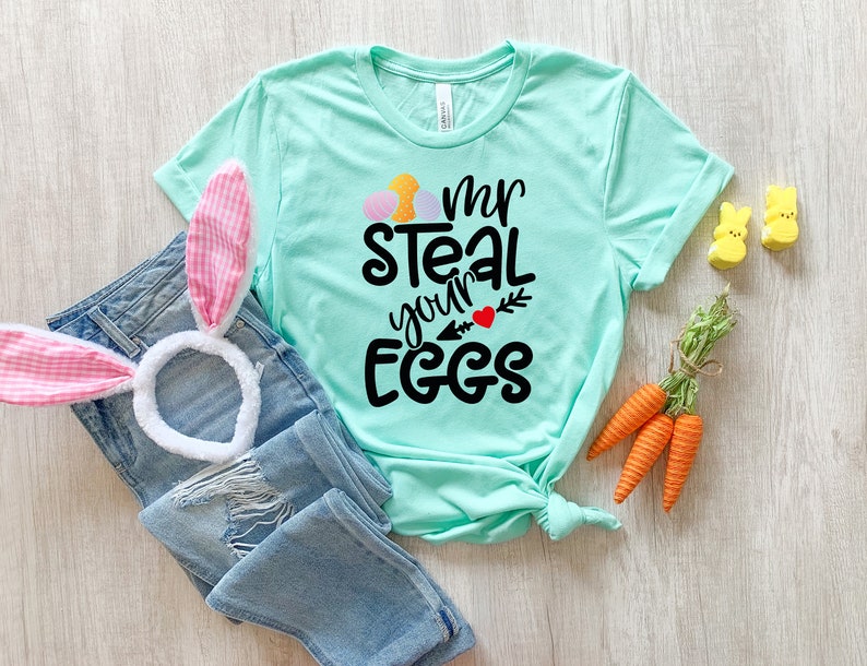 May include: A turquoise blue t-shirt with the text "I'm steal your eggs" in black lettering with a red heart and a green arrow. The shirt is folded and laid on a wooden surface with a pink bunny ear headband, carrots, and yellow chick figurines.