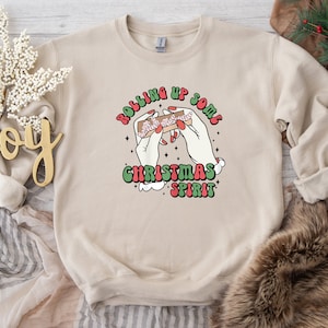 Puede incluir: Una sudadera beige con un estampado gr&aacute;fico de una mano enrollando un porro con el texto "Rolling Up Some Christmas Spirit" en una fuente retro.