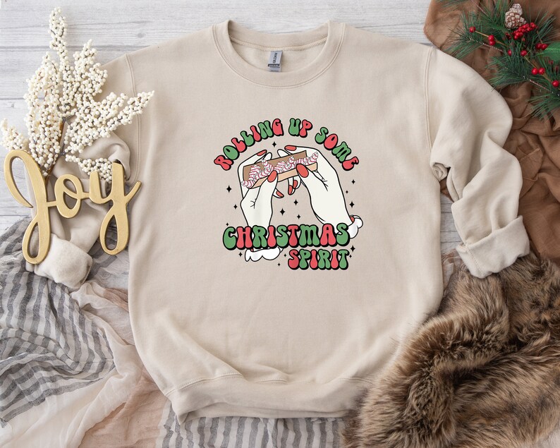 Puede incluir: Una sudadera beige con un estampado gr&aacute;fico de una mano enrollando un porro con el texto "Rolling Up Some Christmas Spirit" en una fuente retro.
