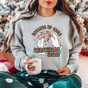 Puede incluir: Una sudadera gris con un gr&aacute;fico de manos enrollando un porro con el texto "Rolling up some Christmas spirit".
