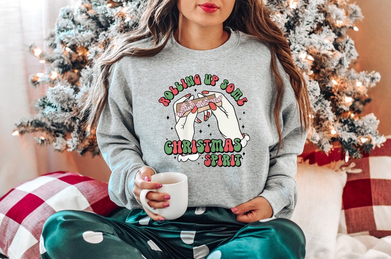 Puede incluir: Una sudadera gris con un gr&aacute;fico de manos enrollando un porro con el texto "Rolling up some Christmas spirit".