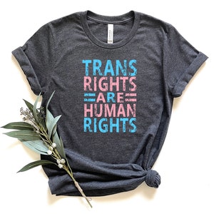 May include: A dark heather grey t-shirt with the text "TRANS RIGHTS ARE HUMAN RIGHTS" printed in pink and blue.